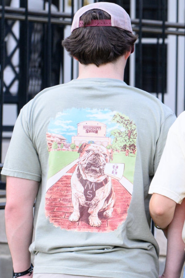 Mississippi State Tan T-Shirt with an image of Bully the bulldog holding a cowbell in it’s mouth on the brick walk into campus