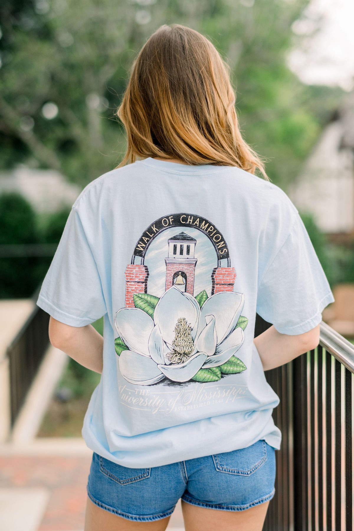 Ole Miss Light Blue T-Shirt with a large image of a magnolia flower in front of the walk of champions gate