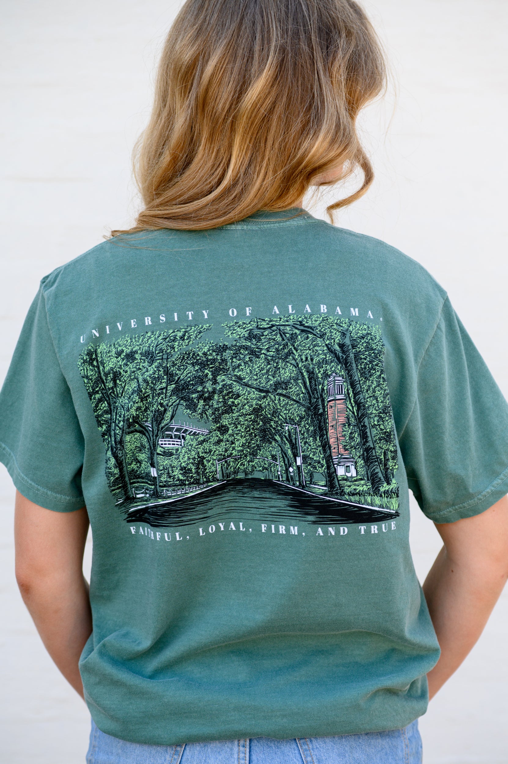 Person wearing a green t-shirt with a graphic design on the back, featuring text that reads "University of Alabama" on the top and "Faithful, Loyal, Firm, and True" on the bottom and a anddrawn landscape illustration of University Boulevard showing Denny Chimes and Bryant-Denny Stadium.